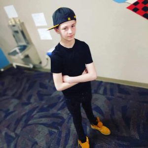 Young Caucasian male stands with his arms crossed, wearing a sideways blue cap, black tshirt, black pants and tan shoes