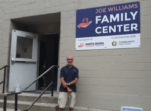 Man in blue shirt nd tan shorts standing in front of a beige brick wall and a sign that says Joe Williams Family Center